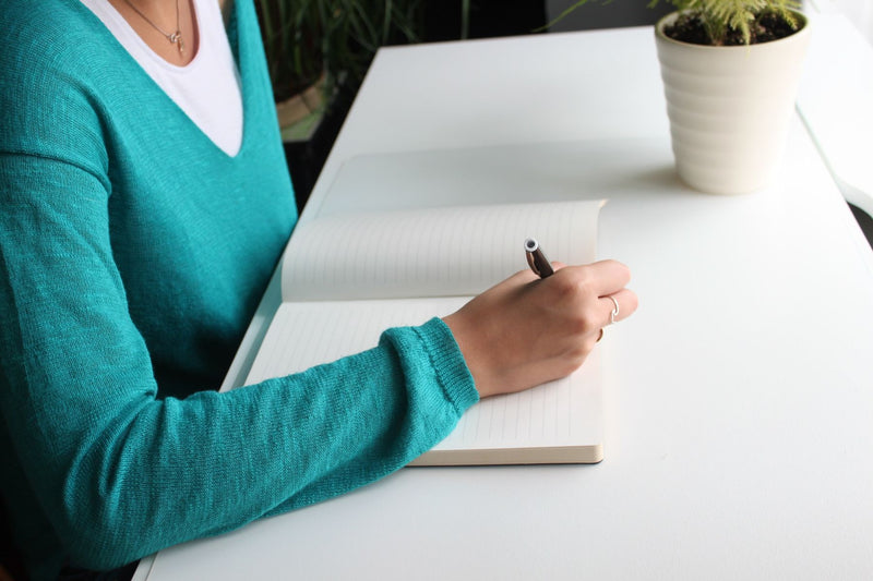 Woman writing in a notebook at a bright, minimal white desk with a plant