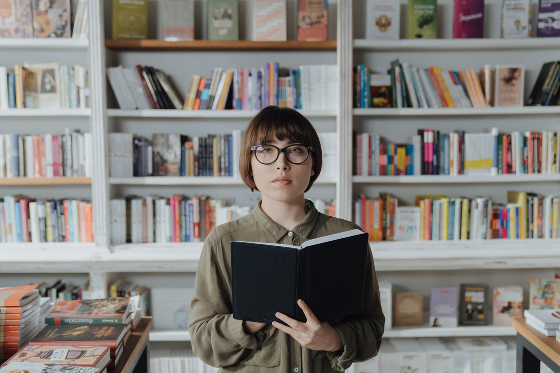 Young woman in glasses reading a book among the shelves of a bookshop