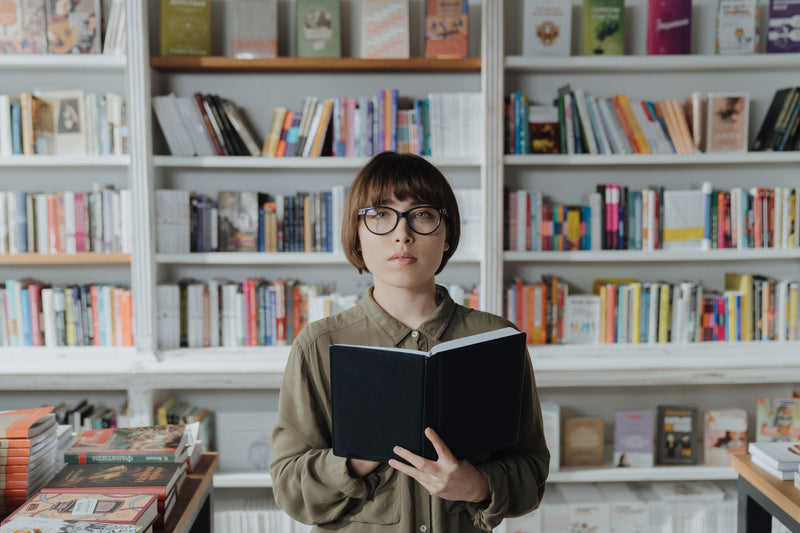 Young woman in glasses reading a book among the shelves of a bookshop