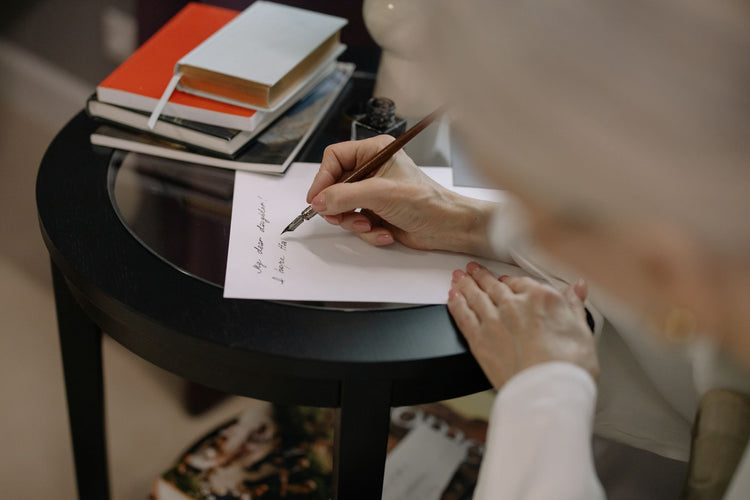 Person writing on white paper with a fountain pen beside stacked books