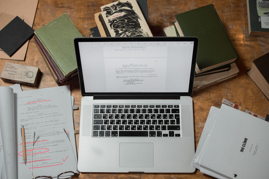 Laptop surrounded by printed manuscript pages and books on a sunlit wooden desk