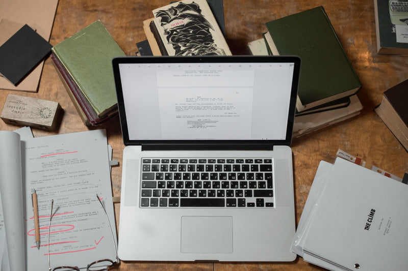 Laptop surrounded by printed manuscript pages and books on a sunlit wooden desk