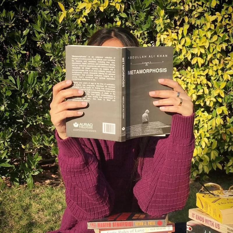 Person reading a book titled 'Metamorphosis' with stacks of books in the foreground and greenery in the background